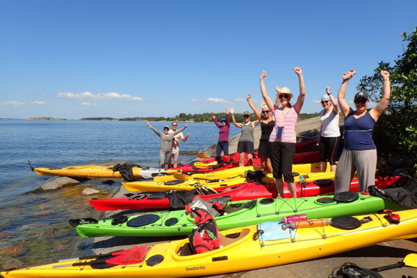 A large group of paddlers by their kayak with their arms raised to the sky.
